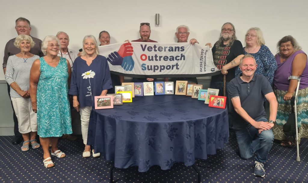 A group of people standing behind a round table. On the table is a collection of framed photos. The people directly behind the table are holding up a banner with the Veterans Outreach Support logo on it. | VOS