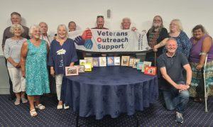 A group of people standing behind a round table. On the table is a collection of framed photos. The people directly behind the table are holding up a banner with the Veterans Outreach Support logo on it. | VOS