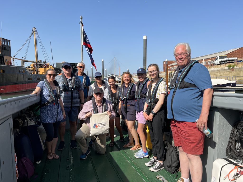 A group of people standing on an LCVP vessel, with one person in the middle kneeling down holding a tote bag with the Veterans Outreach Support logo on it | VOS