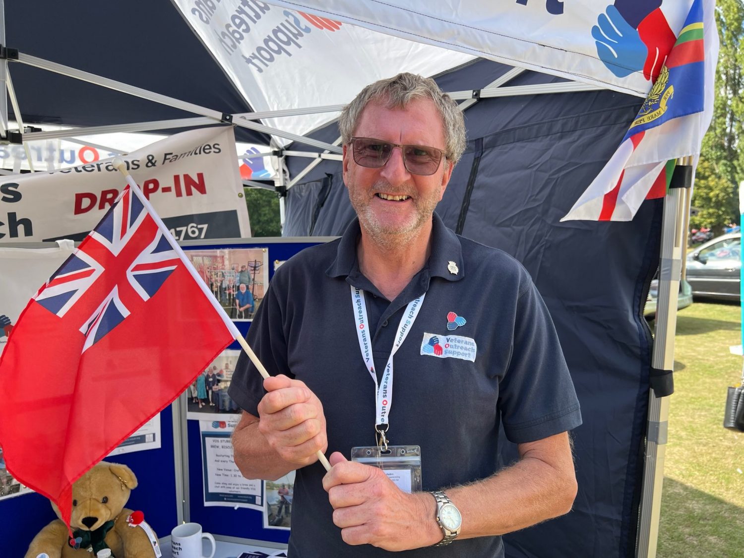A person wearing a navy blue polo with the Veterans Outreach Support charity logo on it, a Veterans Outreach Support lanyard on, holding a Merchant Navy Red Ensign flag | VOS