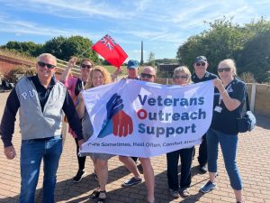 A group of people holding a white banner with the Veterans Outreach Support charity logo on it. One of them is holding a small Red Ensign, which is a red flag with the Union Jack in the top left corner. | VOS
