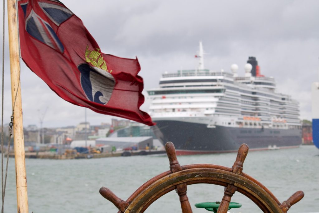 A view of an old Red Ensign flag on the back of the ship, with the wooden wheel at the bottom. In the background is a large cruise ship. | VOS