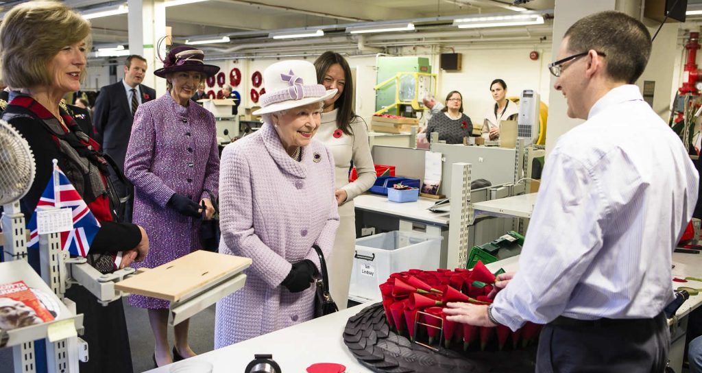 Her Majesty Queen Elizabeth II visiting The Poppy Factory. She is wearing a lilac coat and had, and is speaking with an employee who is showing her a Remembrance wreath. | VOS