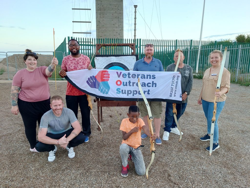 A group of people standing around an archery target holding bows and arrows. Two of them are holding up a white banner with the Veterans Outreach Support charity logo on it. | VOS