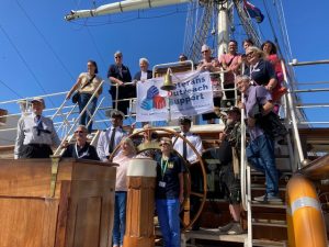 A group of people aboard RNOV Shabab Oman II. They are standing around the wheel, with some on the same level as the wheel, others on steps upwards and the rest on the level above the wheel. The people on the upper level are holding a white banner with the Veterans Outreach Support logo on it. They are holding it against the railings on their level and it is hanging down below the ship's bell. | VOS