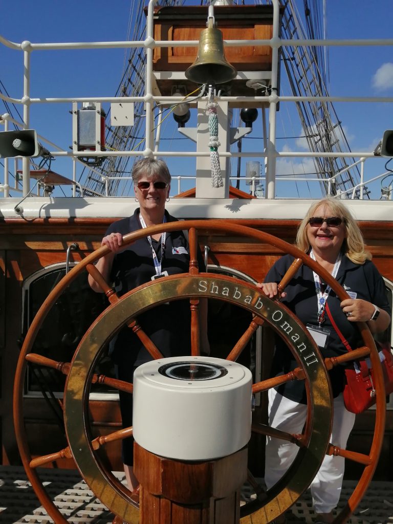 Two people wearing navy polo shirts with the Veterans Outreach Support charity logo on them, standing behind the large wooden wheel of RNOV Shabab Oman II. The wheel has a gold strip running around the inner wooden section which is engraved with the ship's name. Above them is the golden ship's bell. | VOS