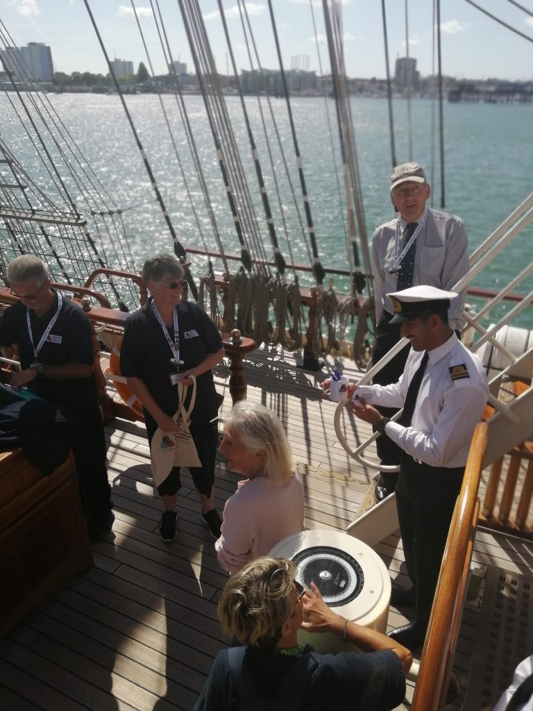A view from above of six people on RNOV Shabab Oman II. Two of them are wearing navy polo shirts with the Veterans Outreach Support charity logo on them, and they have just given a member of the ship's crew a selection of gifts with the same charity logo. The crew member is wearing black trousers, a white shirt, a black tie and a black and white navy cap. In the background is Portsmouth Harbour water and Gosport. | VOS