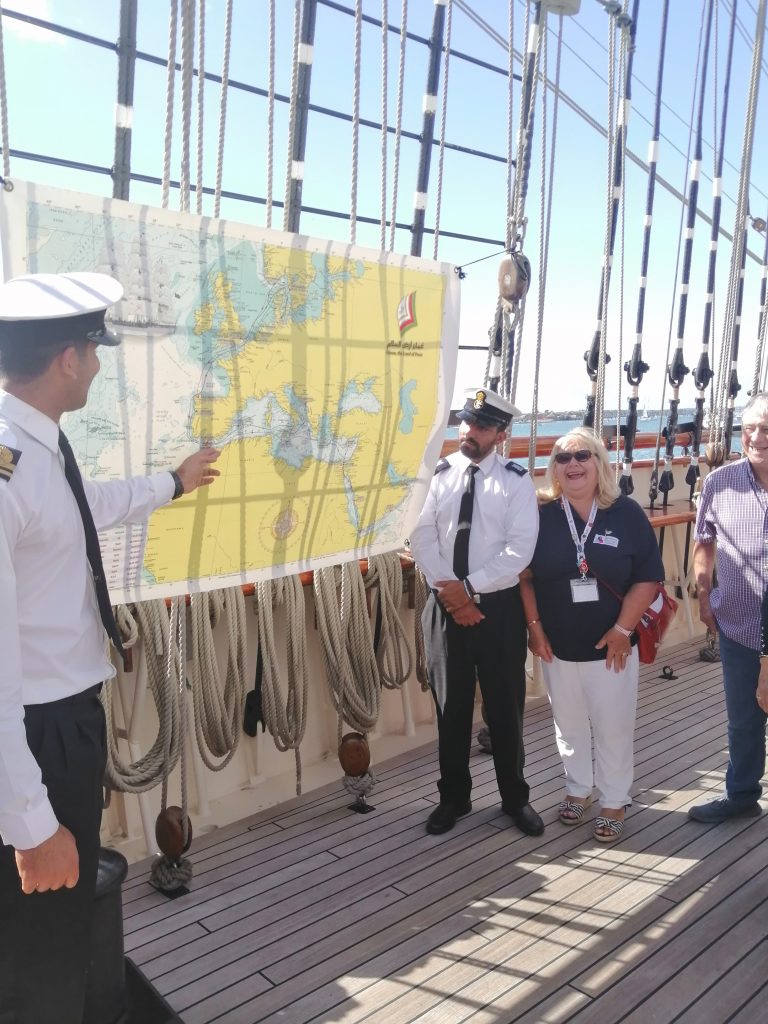 Two crew members aboard RNOV Shabab Oman II pointing at a navigational map which has been hung up from the rigging at the side of the ship. Next to them is a person wearing a navy polo shirt with the Veterans Outreach Support logo on it, who is smiling at the camera. | VOS