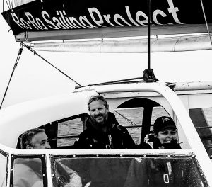 A black and white photo of three smiling people in the cockpit of a sailing yacht, viewed from outside the cockpit. | VOS