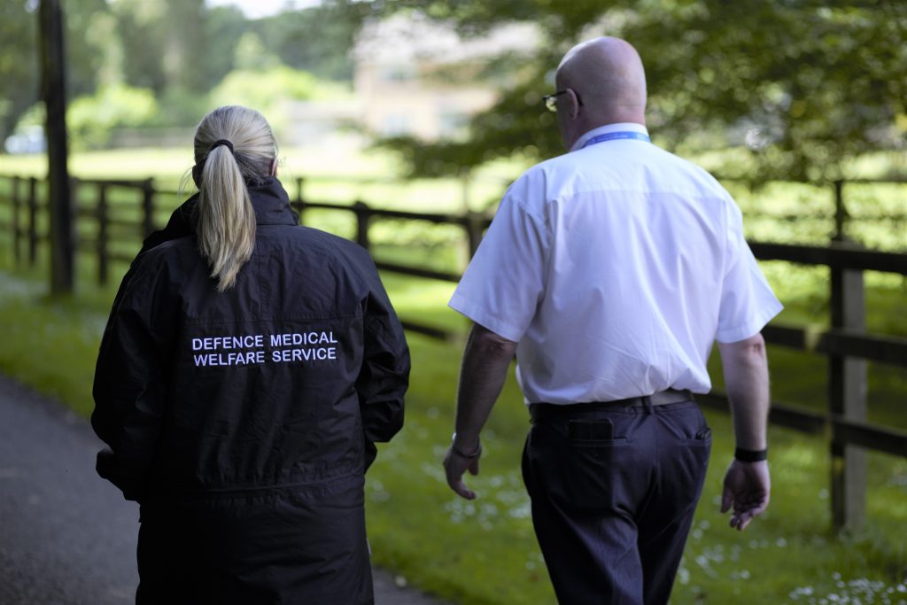 Two people walking along on a path next to a fenced off field with trees on it. The person on the right is wearing a white shirt, and the person on the left is wearing a long black coat with the words "Defence Medical Welfare Service" on it in white. | VOS