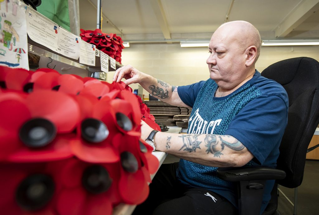 A person with tattoos on their arm, who is sitting at a desk which has Remembrance wreaths made of plastic poppy flowers on it. | VOS