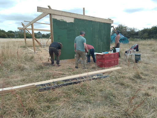 Four people building a wooden structure in a field. They are painting the end wall green. | VOS