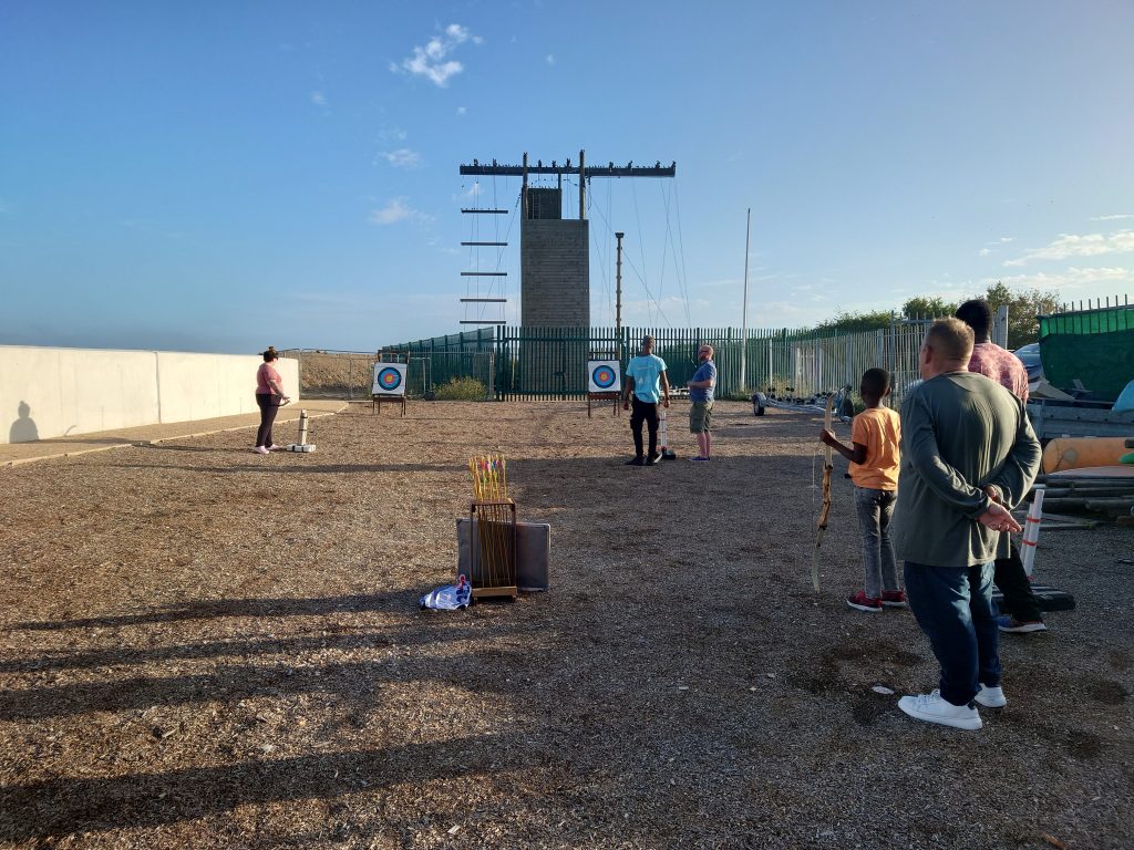 Two people are preparing archery bows to fire arrows at targets. A group of other people are standing in the foreground watching. In the background is a climbing wall and behind it the sky is blue with an evening sunset. | VOS