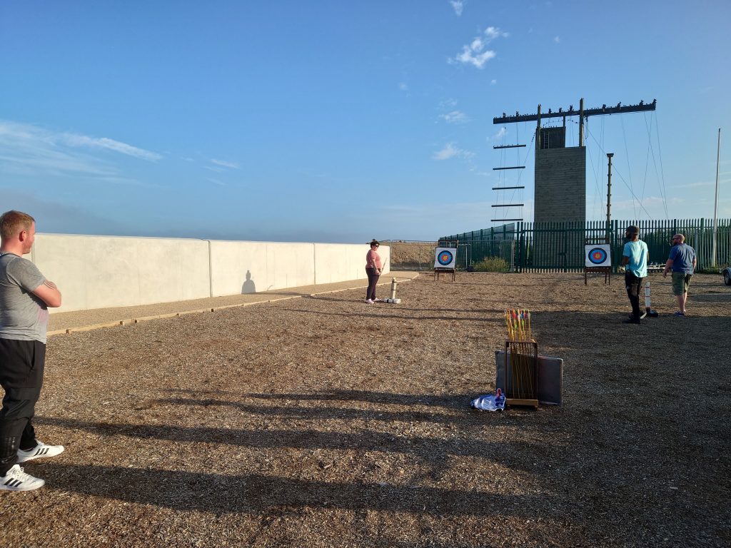 Two people are preparing archery bows to fire arrows at targets. One person is standing to the left in the foreground watching. In the background is a climbing wall and behind it the sky is blue with an evening sunset. | VOS