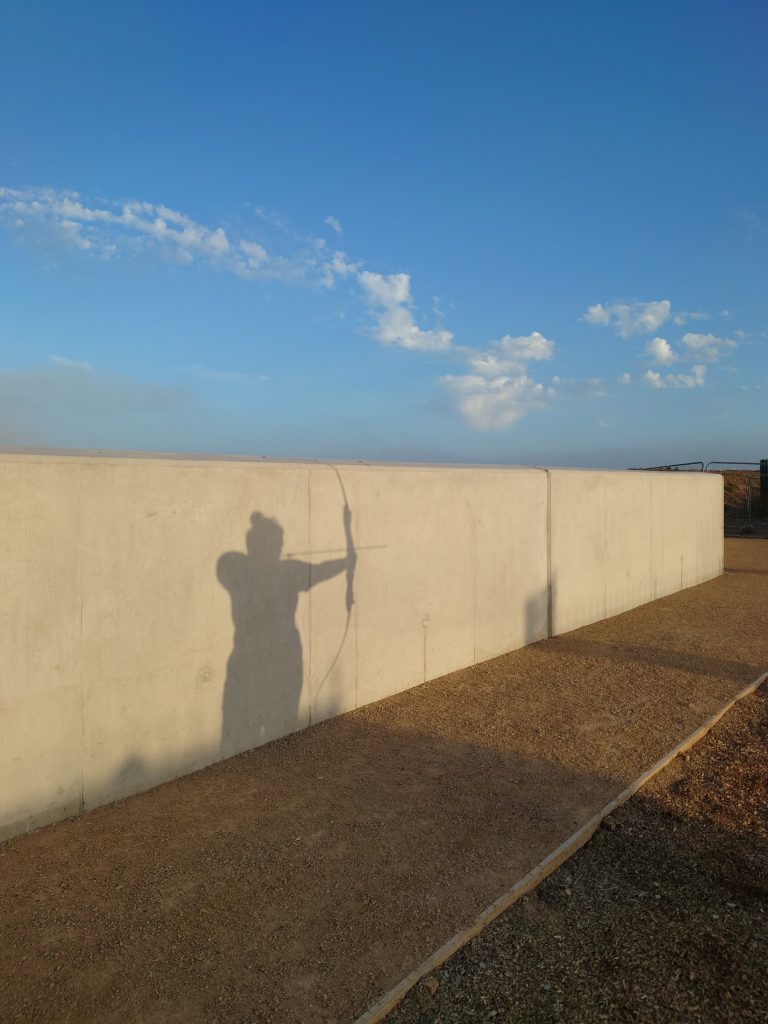 A picture of a short white wall with a shadow of a person standing holding an archery bow ready to fire an arrow. In the background the sky is blue with a summer evening sunset. | VOS