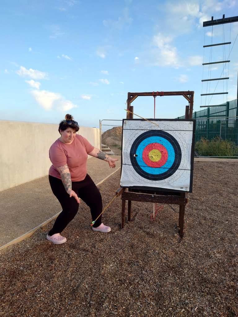 A person posing next to an archery target, pointing at the arrows they've fired which have missed the target. In the background the sky is a summer evening sunset. | VOS