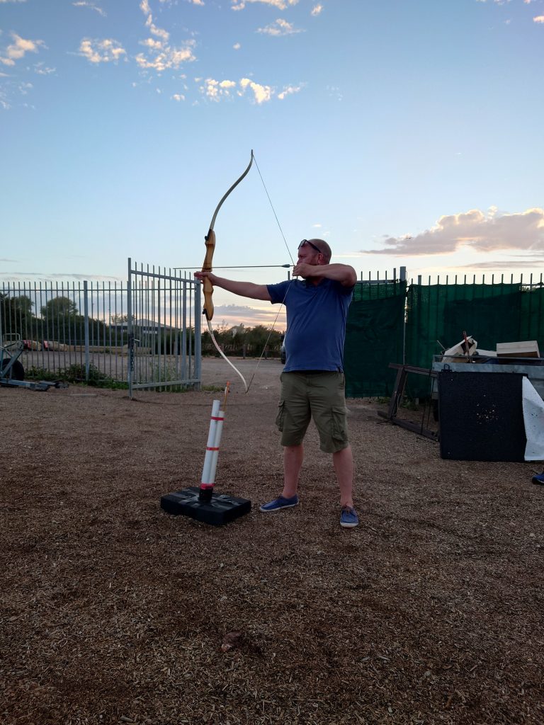 A person standing holding an archery bow, ready to fire an arrow at a target which isn't visible. In the background the sky is a nice summer evening. | VOS