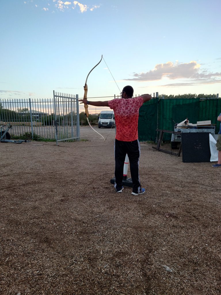 A person standing holding an archery bow, ready to fire an arrow at a target which isn't visible. In the background the sky is a nice summer evening. | VOS