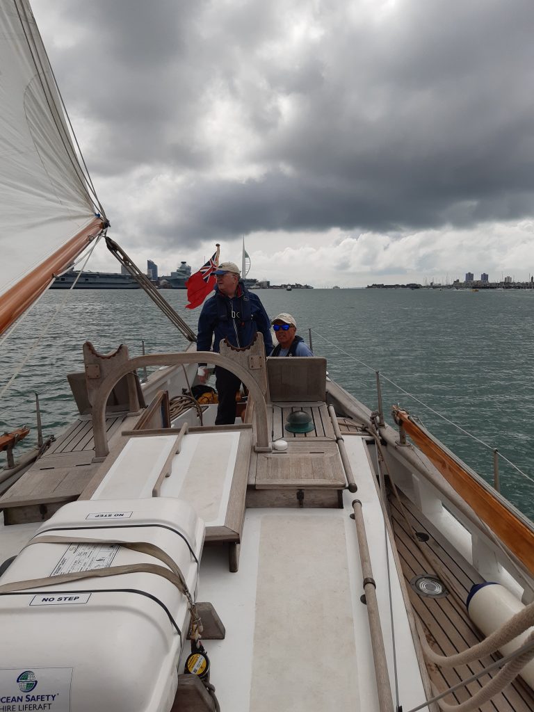 A view of two people in the cockpit of junk yacht Boleh, sailing in Portsmouth Harbour. Some of the mainsail is visible to the left. The sky behind them is great and cloudy and the sea is somewhat great too. | VOS