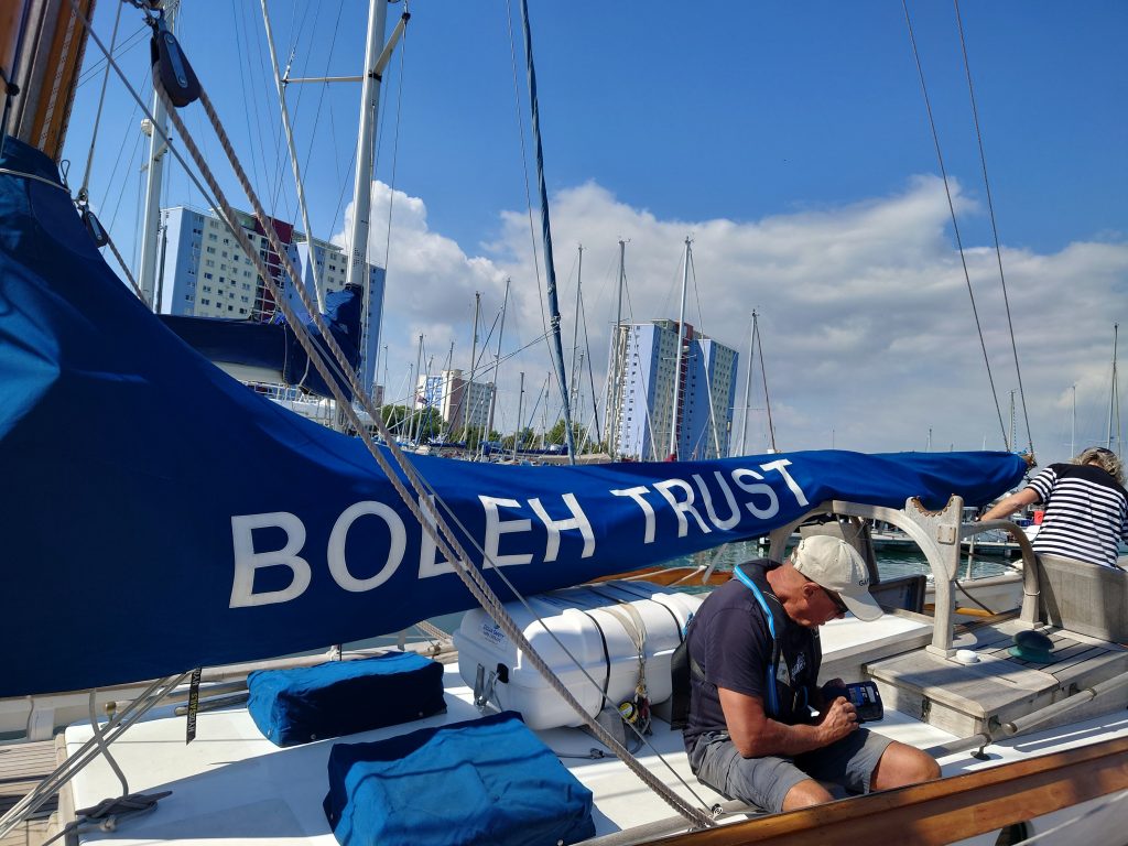A photo of junk yacht Boleh in a marina with a blue cover on the mainsail which has the words "Boleh Trust" on it. In the background the sky is bright blue with some white clouds. | VOS
