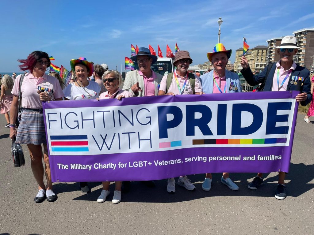 A group of people standing in a line holding up a purple banner with the charity logo of Fighting With Pride (FWP) on it, with the words "The Military Charity for LGBT+ Veterans, seeing personnel and families" on it. The people are also holding or wearing various items with rainbow pride flags on them. The sky is bright blue behind them. | VOS