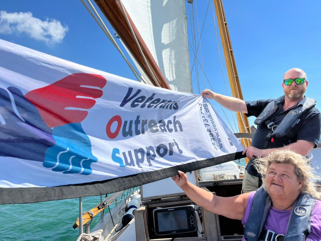Two people holding up one end of a narrow white banner with the Veterans Outreach Support charity logo on it. They are on a sailing yacht called Boleh and part of the yacht and its mainsail are visible. In the background some sea is visible and the sky is bright blue. | VOS