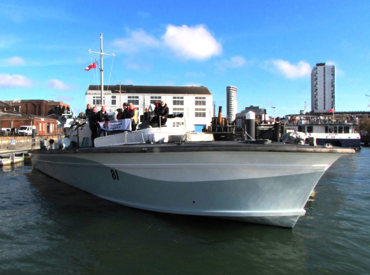 A photo of Motor Gun Boat 81, which is a small Royal Navy vessel from World War II. She is moored outside Boathouse 4 in Portsmouth Historic Dockyard. There's a group of people aboard her holding up a white banner with the Veterans Outreach Support charity logo on it. | VOS