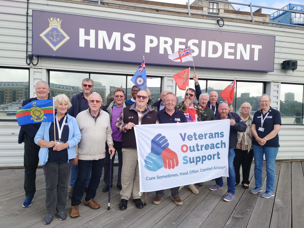 A group of people standing outside HMS President in London. At the front in the middle two people are holding up a white banner with the Veterans Outreach Support charity logo on it. | VOS