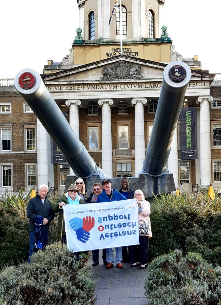 A photo of a group of people standing in front of a statue of two large military guns outside the Imperial War Museum in London. Two people are holding up a white banner with the Veterans Outreach Support charity logo on it, but the banner is upside down. | VOS