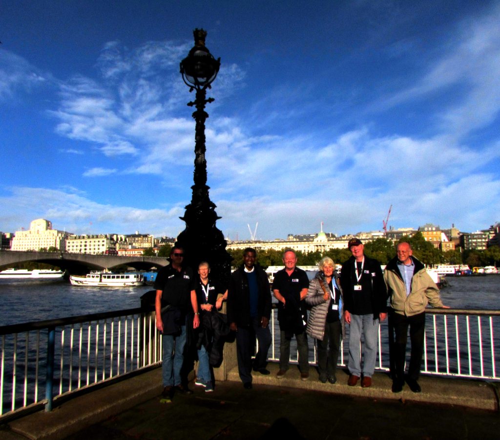 A group of people standing on a path at the side of the River Thames in London. There is an old-fashioned lamppost directly behind them, and in the far background on the other side of the river we can see an array of London buildings. | VOS