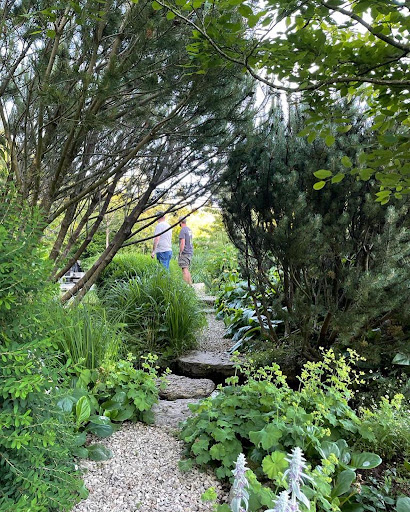 A view of a grey gravel path through a garden with luscious green bushes and trees. In the distance are two people, one of whom is wearing a t-shirt with the Veterans' Growth charity logo on the back of it. | VOS
