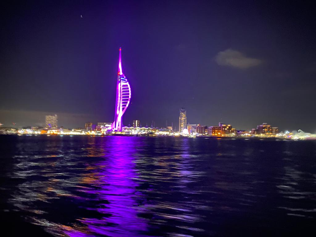 A photo of the Spinnaker Tower lit up in purple and the lights of Gunwharf Quays taken from on the water in Portsmouth Harbour. | VOS