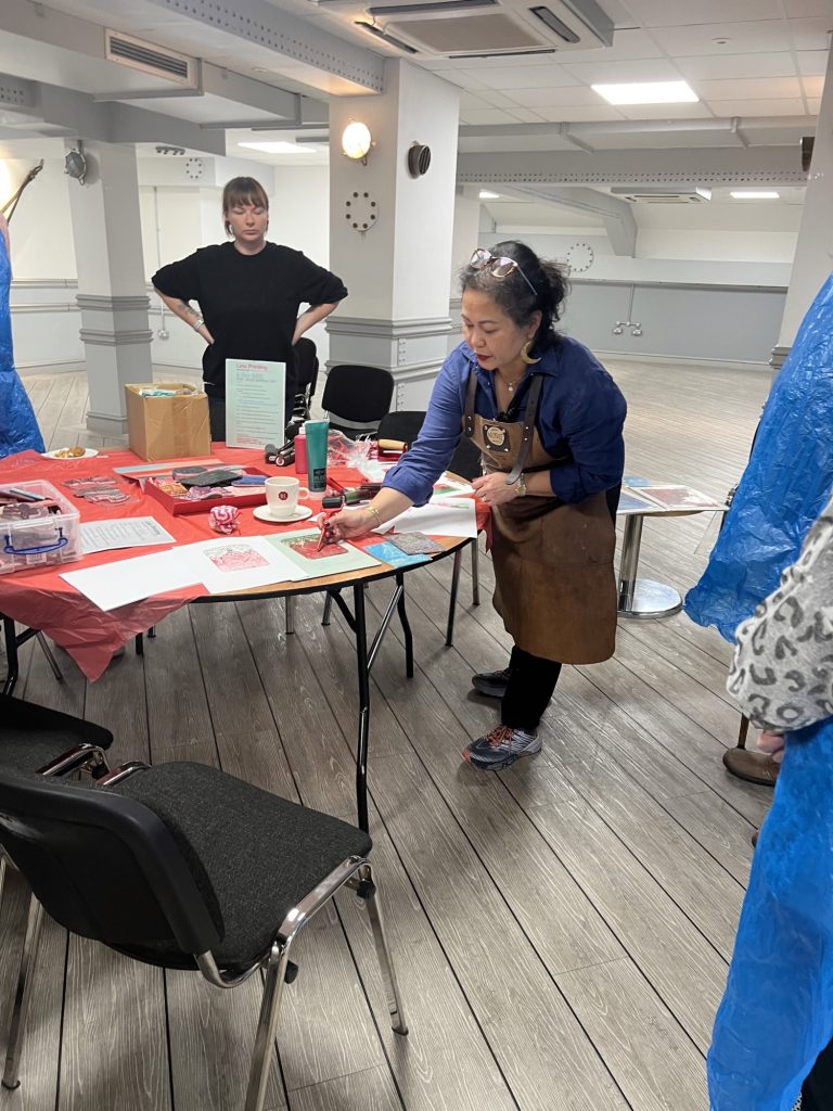 A photo of a person demonstrating how to do lino printing. They are standing at a round table, leaning over to point at something. They are wearing a brown leather apron. | VOS