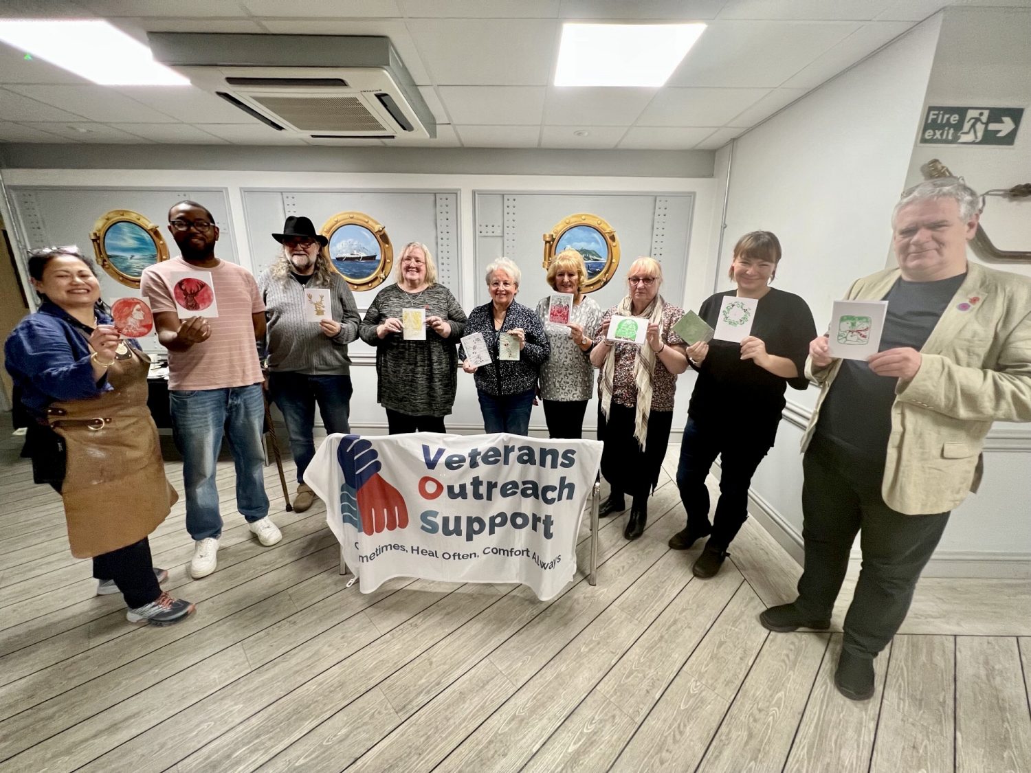 A photo of nine people standing in a semi-circle, each holding up a piece of lino printing artwork that they have made. In front of them, draped over two chairs is a white banner with the Veterans Outreach Support charity logo on it. | VOS