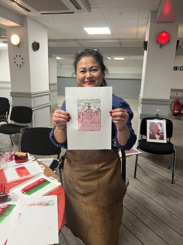A photo of someone holding up a piece of paper with a lino printing design printed on it. They are wearing a brown leather apron and on the table next to them are paints and other lino printing equipment. | VOS